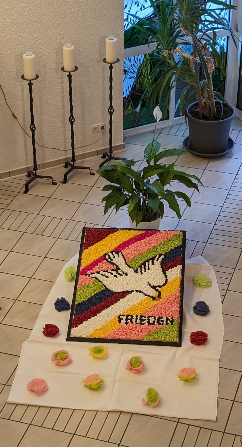 Quadratisches Bild mit einer weißen Taube vor buntem Hintergrund und dem Wort 'FRIEDEN', umgeben von kleinen farbigen Blumen auf einem weißen Tuch, im Hintergrund drei Kerzenständer und zwei große Pflanzen.